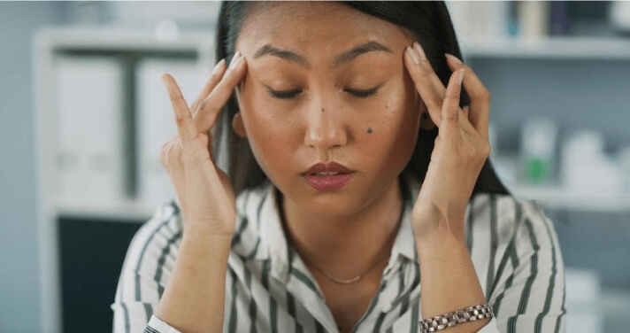 Woman experiencing long COVID symptoms, eyes closed, massaging her forehead.